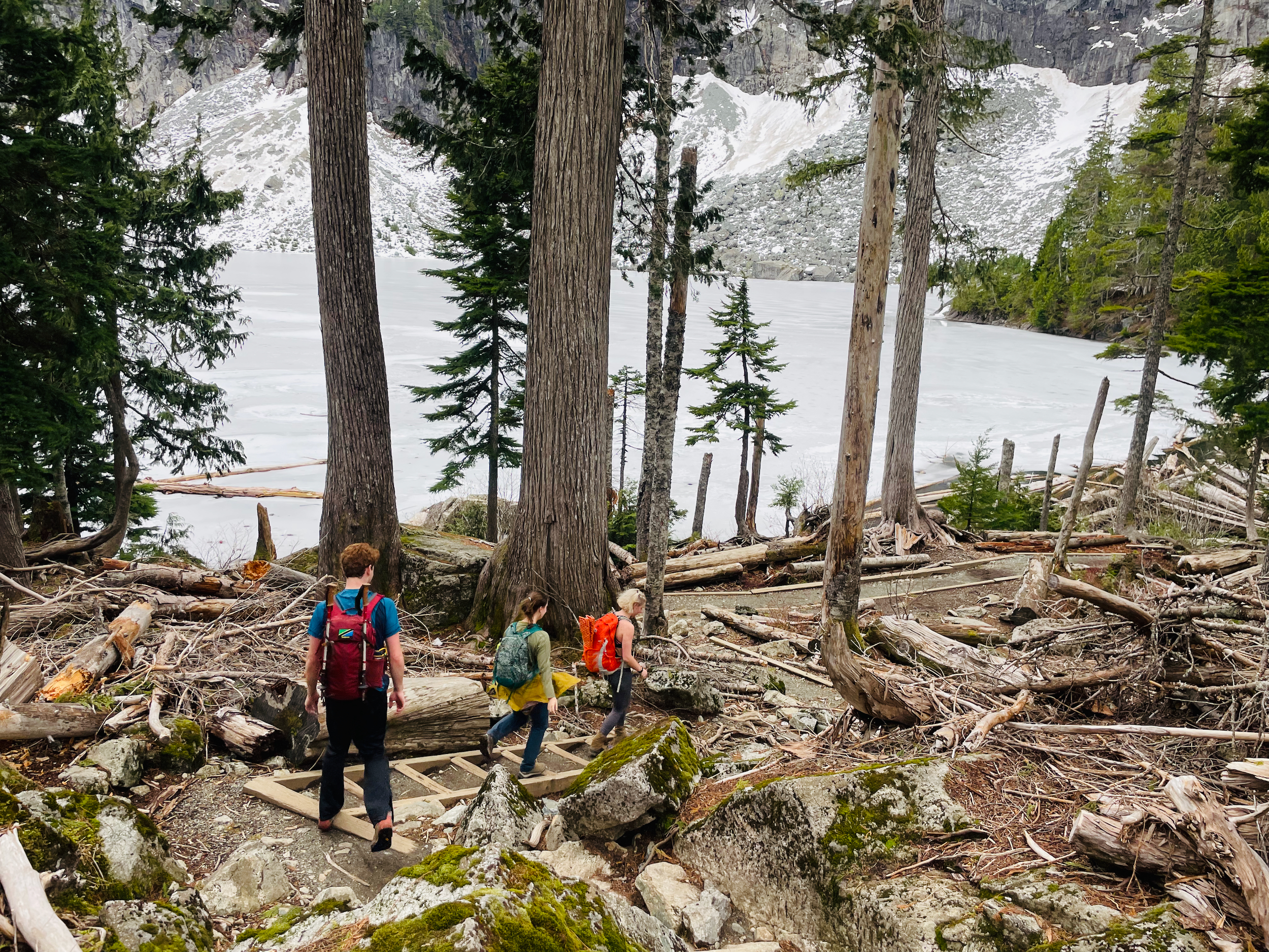 People hiking in the foreground on a trail through trees with an ice covered lake in the background.